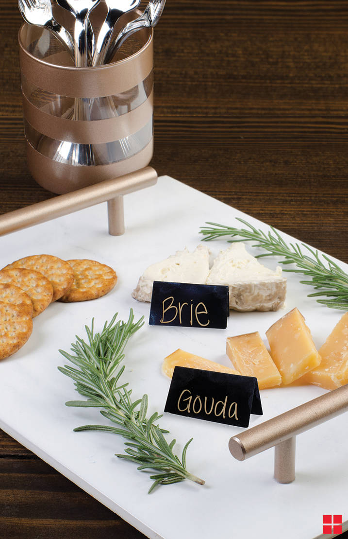 Rose gold handles on a white serving tray that has crackers, pieces of brie and gouda cheese and garnishes on top.